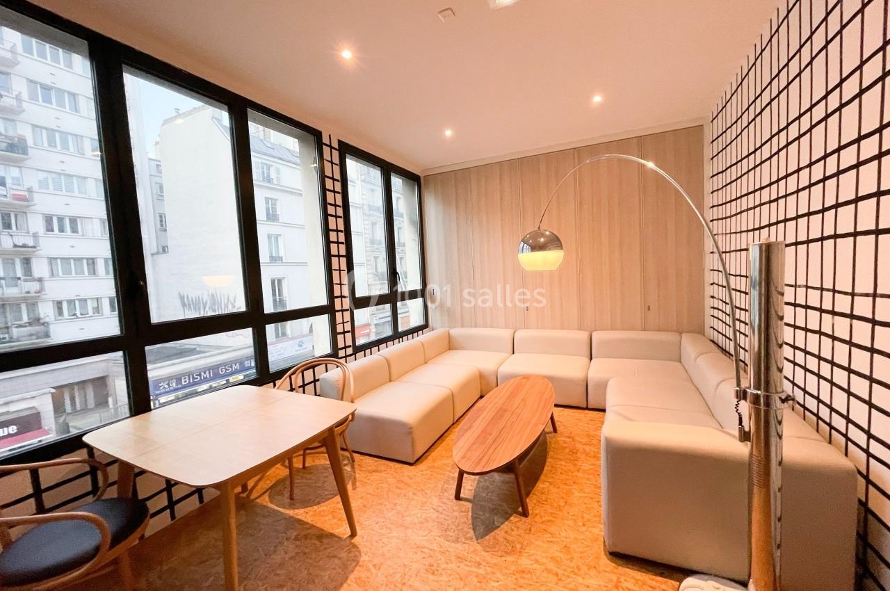 Salle lumineuse avec canapé d'angle beige, table basse en bois, table à manger et grandes fenêtres donnant sur la ville.