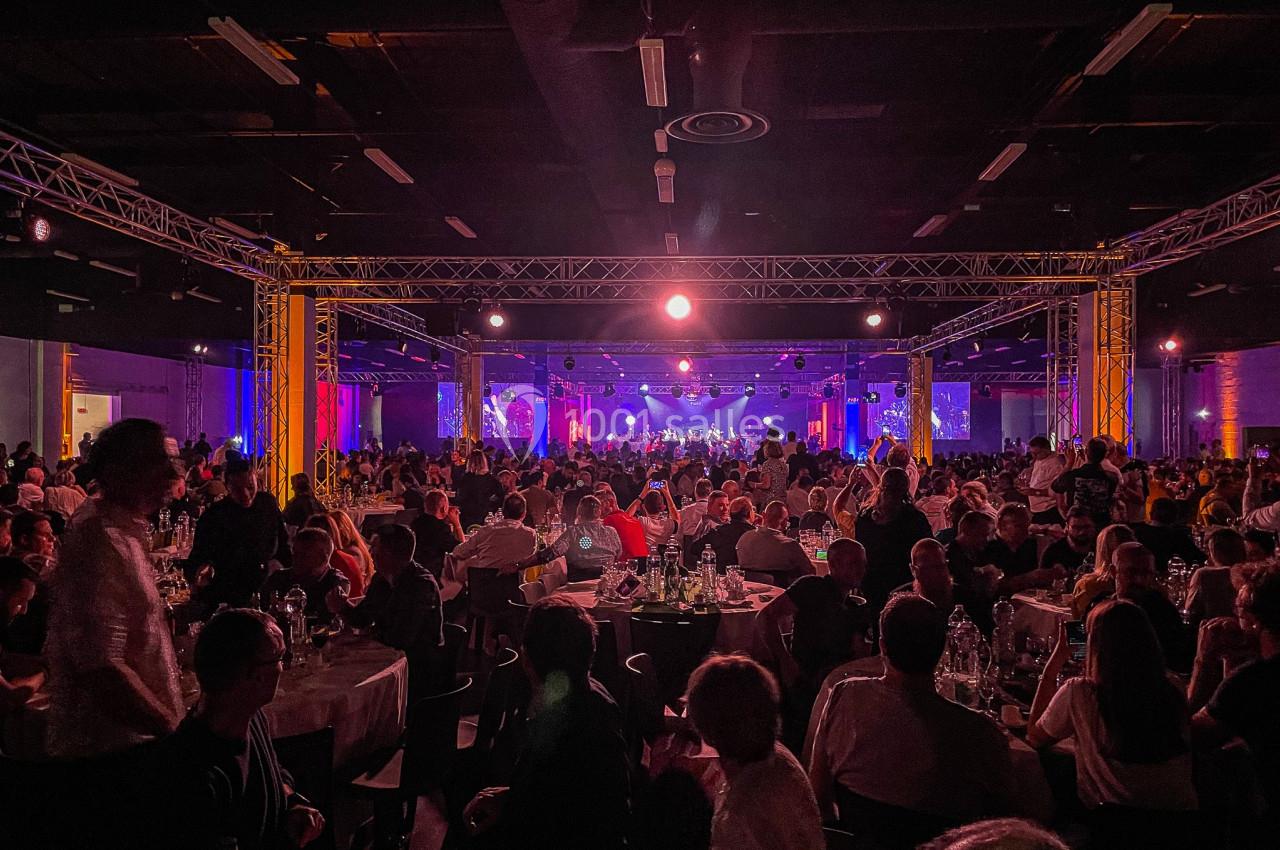 Salle comble avec des tables rondes et une scène éclairée au fond, lors d'un événement en intérieur.
