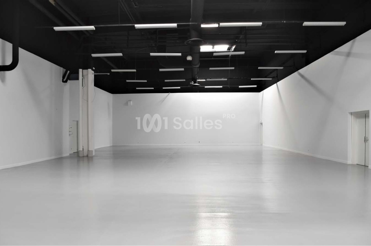 Grand espace intérieur vide avec murs blancs, sol brillant et plafond noir équipé de luminaires suspendus.