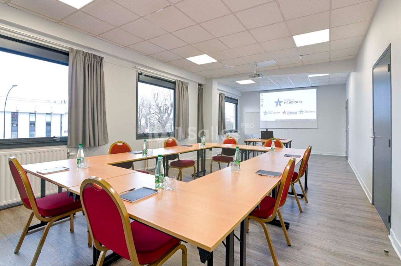 Salle de réunion lumineuse avec tables disposées en U, chaises rouges, bouteilles d'eau et écran de projection.