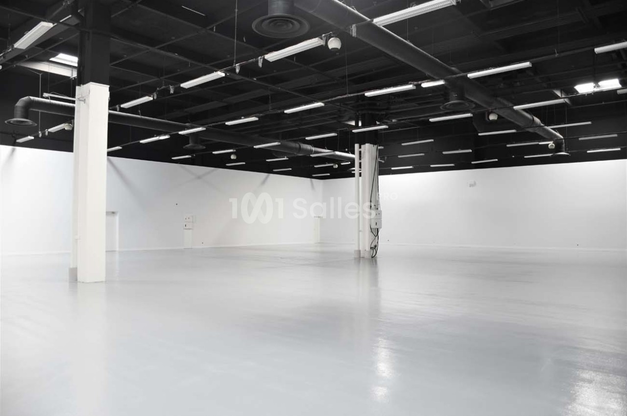 Grand espace intérieur vide avec sol brillant, murs blancs et plafond noir équipé de luminaires industriels.