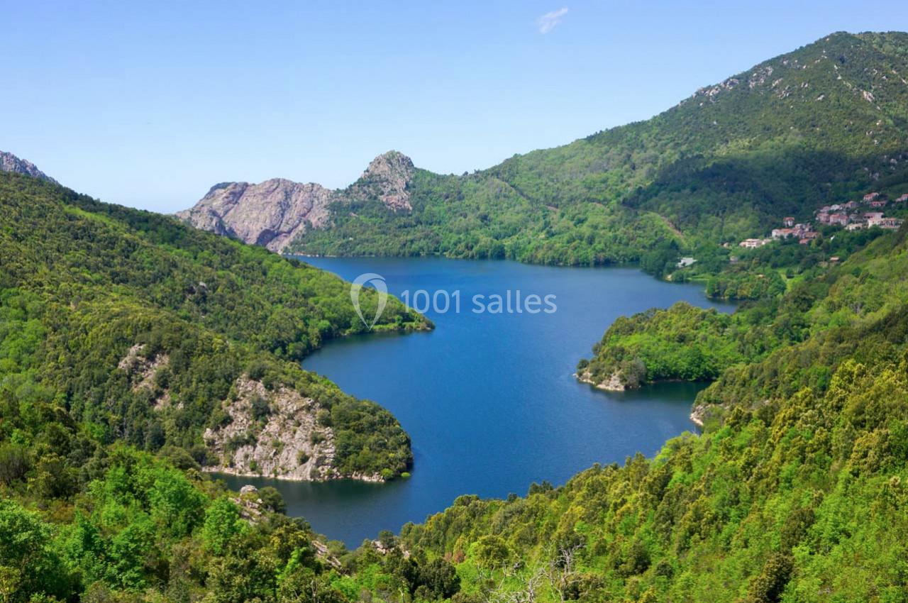 Vue d'un lac entouré de collines verdoyantes et de montagnes sous un ciel bleu clair.