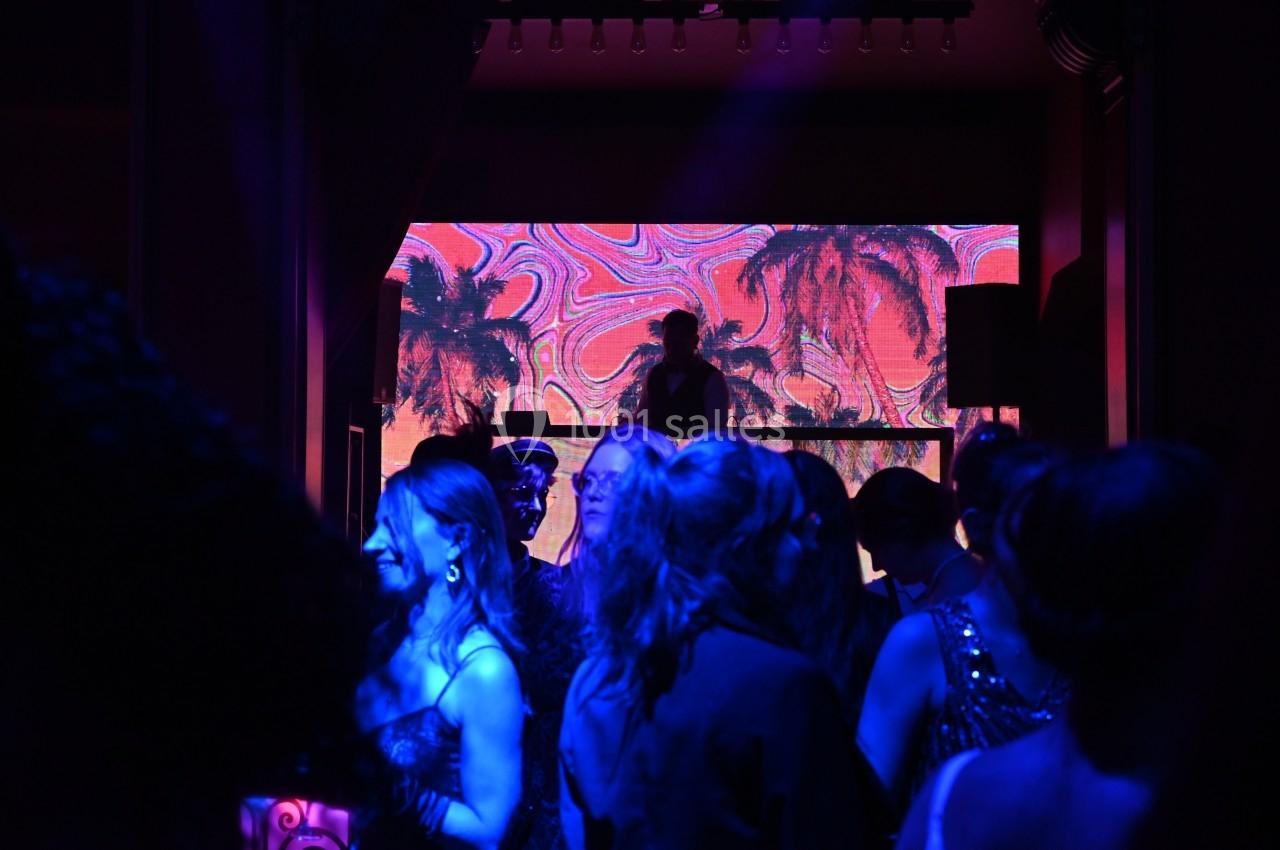 Personnes dansant dans une salle sombre avec un DJ devant un écran lumineux affichant des palmiers colorés.