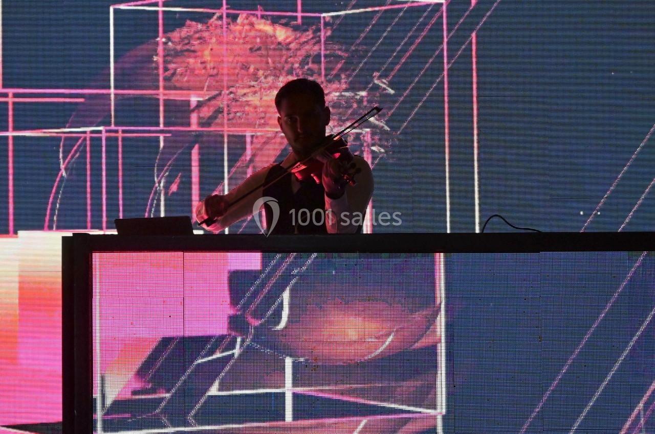 Un musicien joue du violon sur une scène éclairée par des projections lumineuses abstraites en arrière-plan.