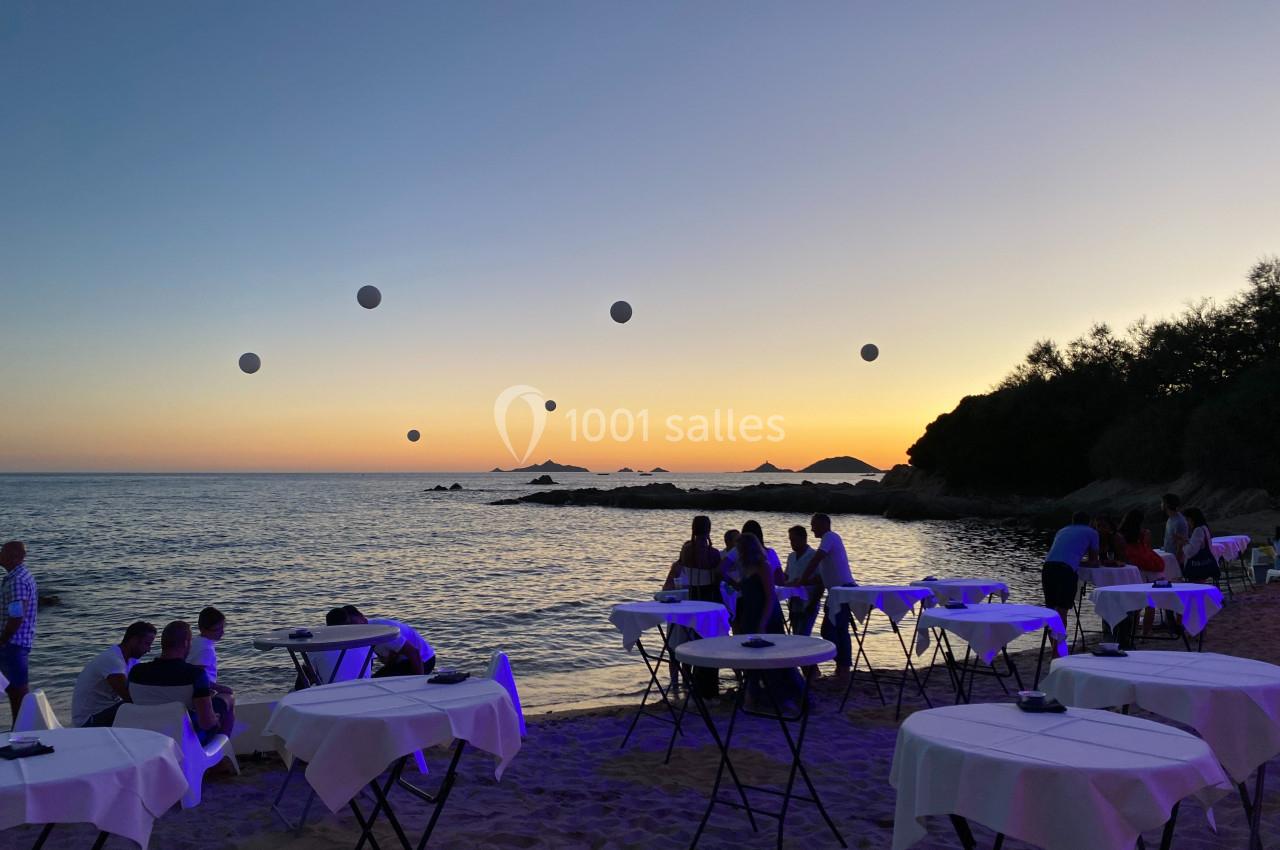 Tables dressées sur une plage au coucher du soleil, avec des invités et des décorations lumineuses flottantes.