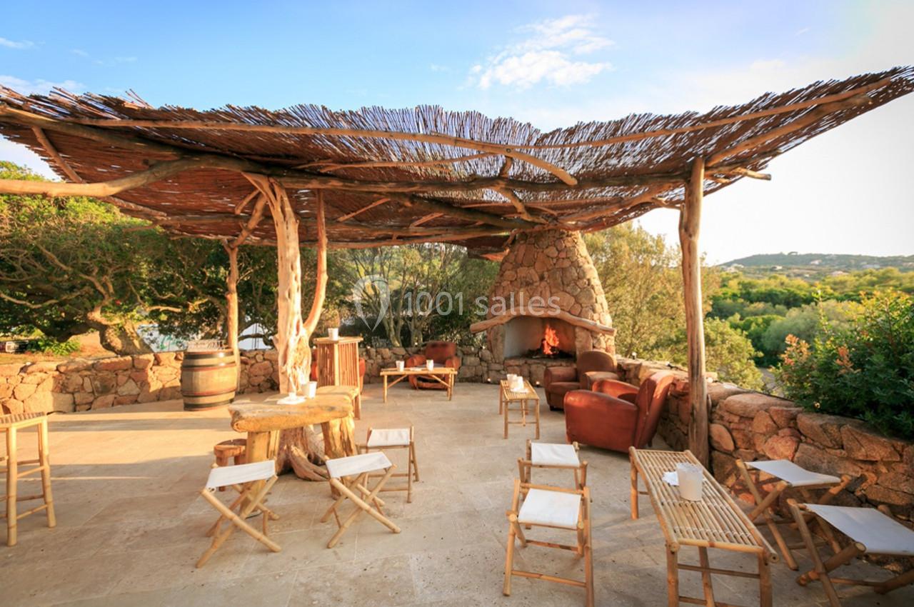 Terrasse en plein air avec mobilier en bois, cheminée en pierre et pergola en branches, entourée de végétation.