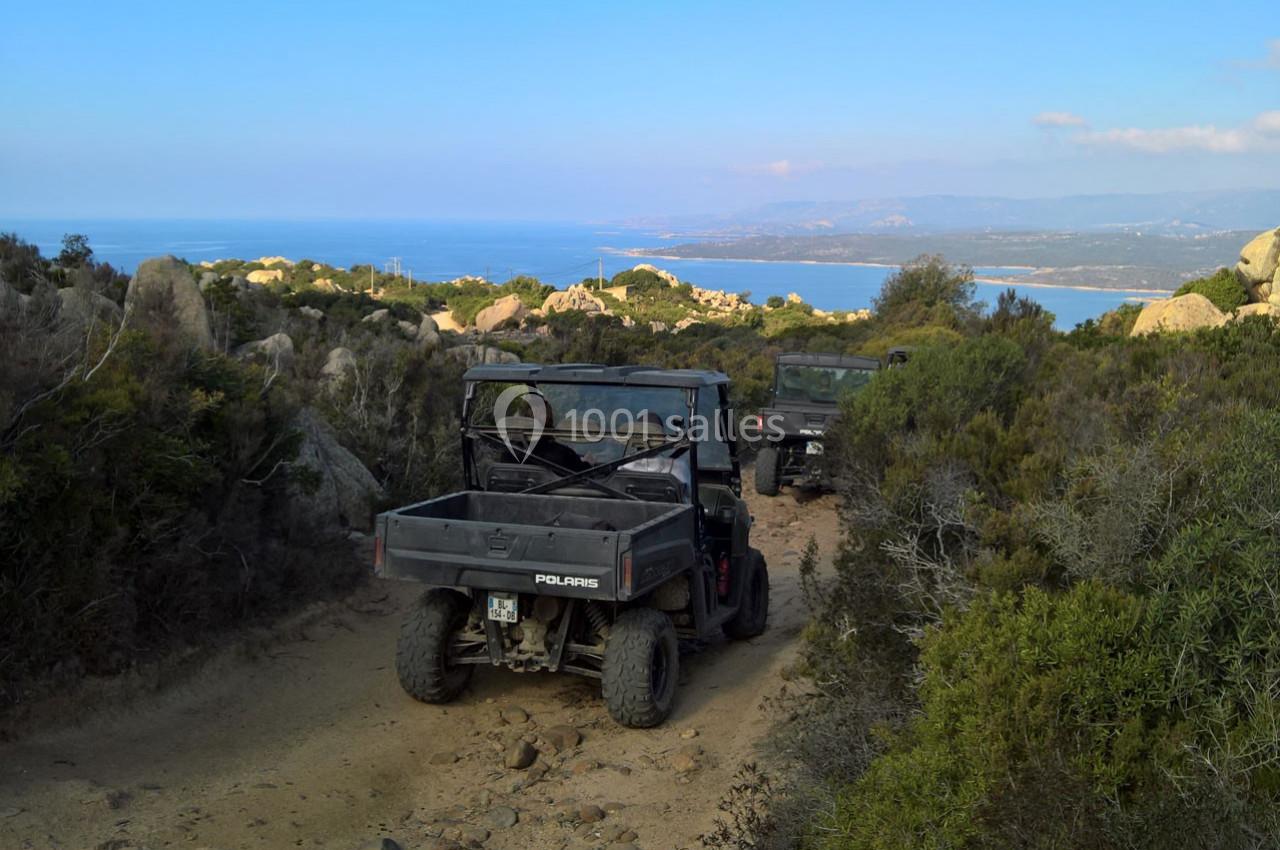 Deux véhicules tout-terrain circulent sur un chemin rocailleux entouré de végétation, avec vue sur la mer au loin.