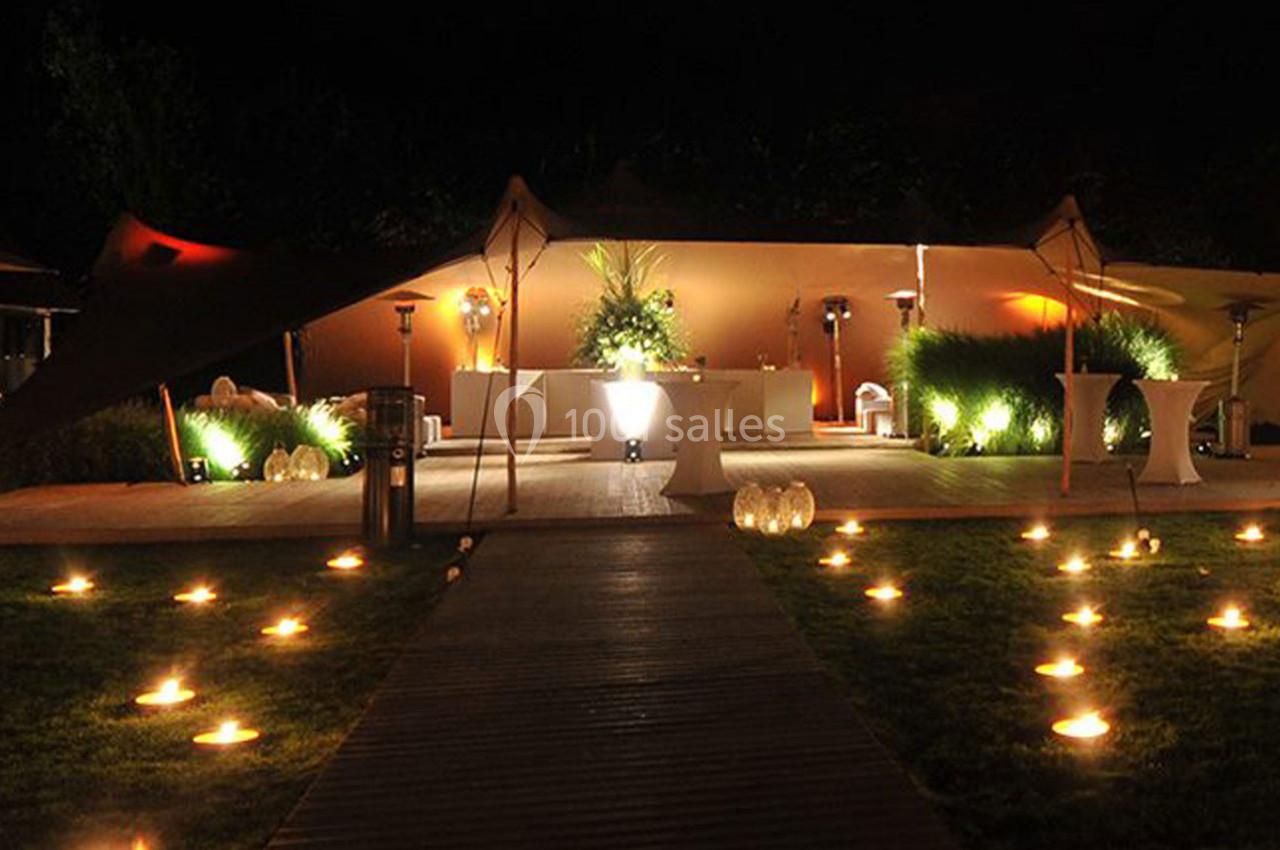 Espace extérieur éclairé de nuit avec des bougies au sol, une tente et une table décorée au centre.