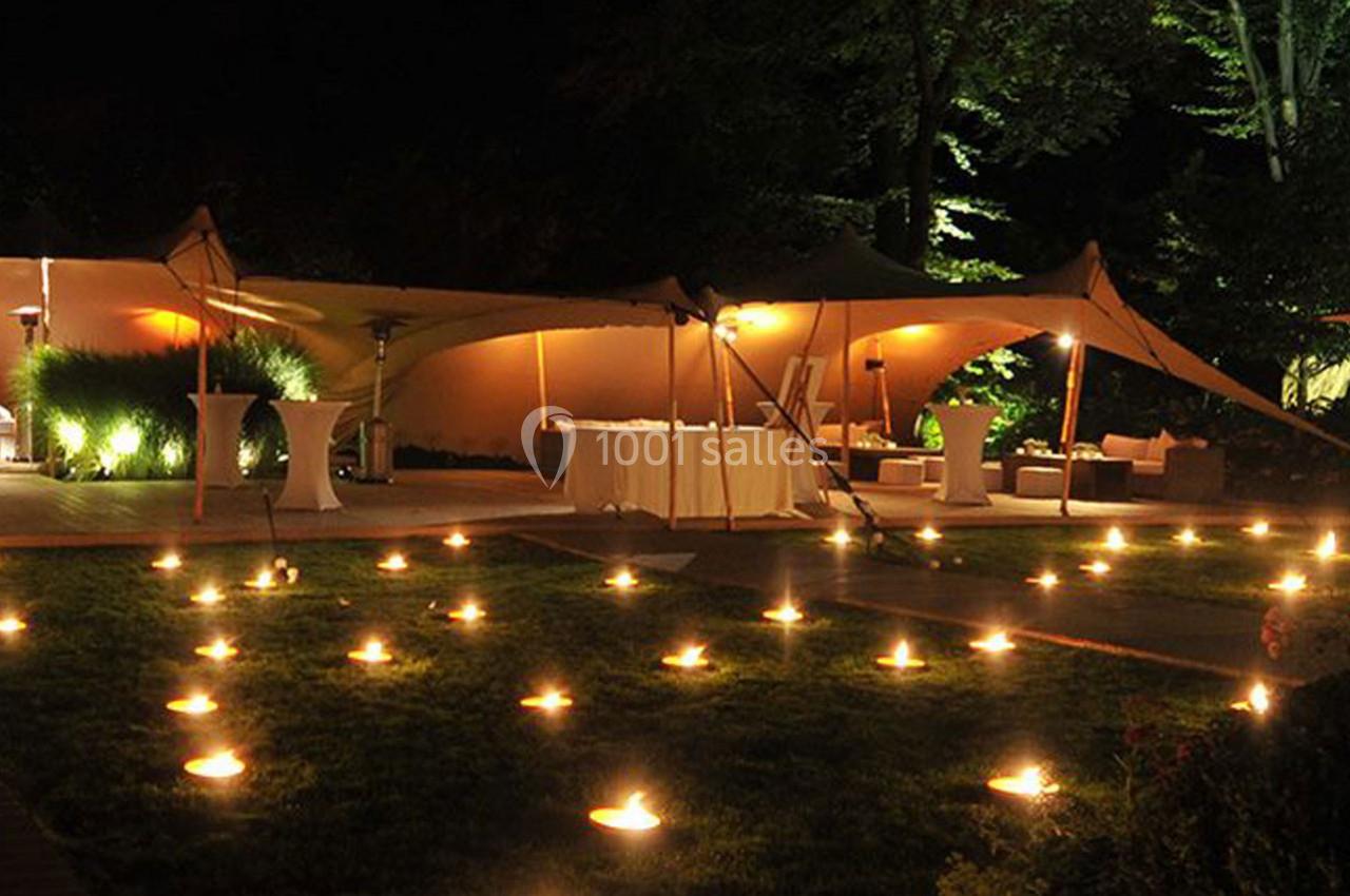 Tente éclairée installée dans un jardin de nuit, entourée de bougies disposées sur la pelouse.