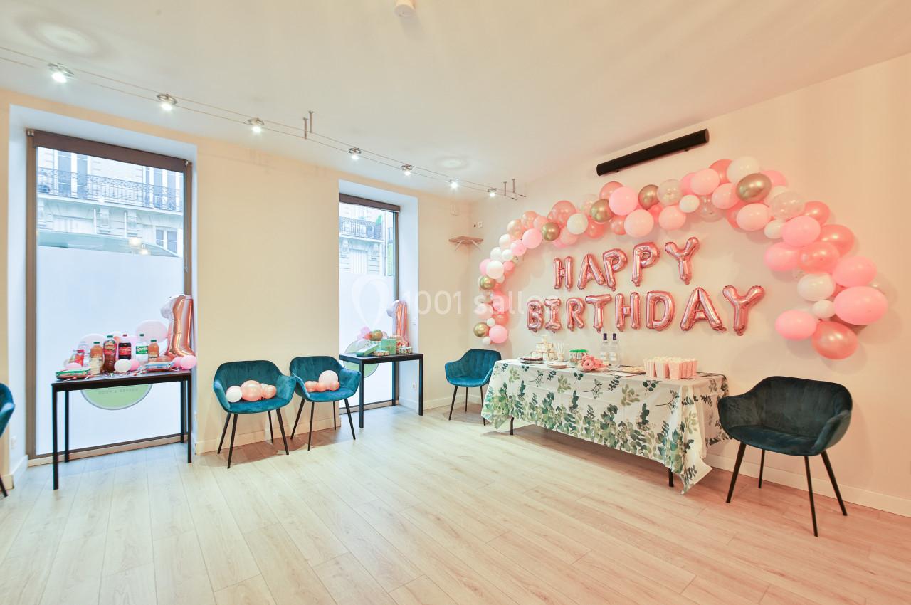 Salle décorée pour un anniversaire avec ballons roses et blancs, table garnie et chaises disposées autour.