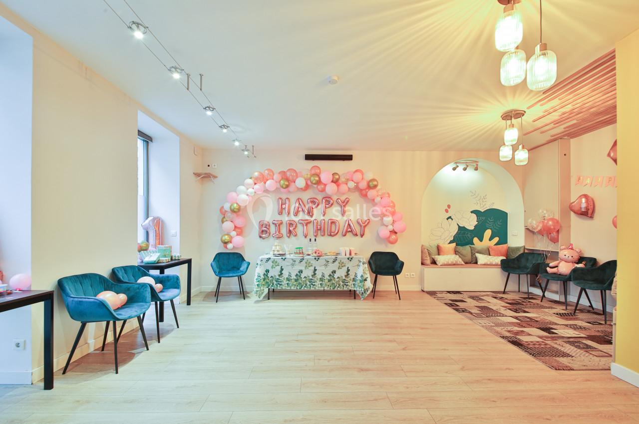 Salle décorée pour un anniversaire avec ballons roses, guirlande ’Happy Birthday’ et table ornée de motifs floraux.