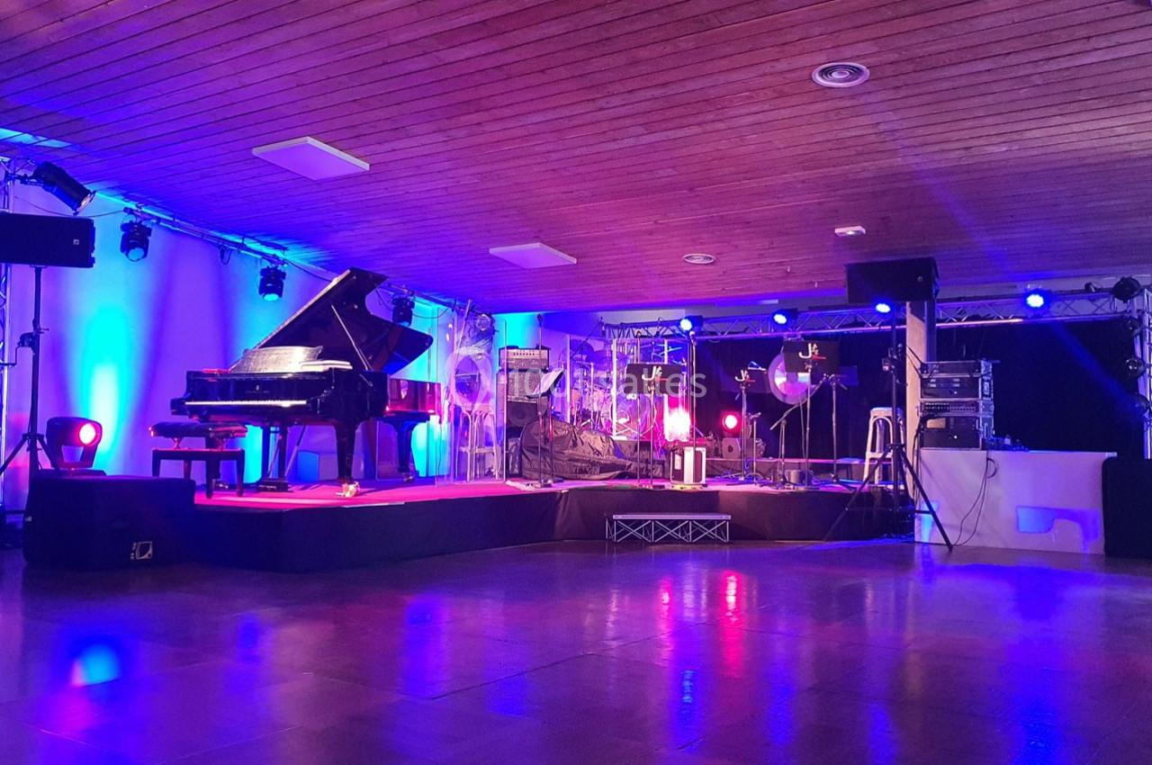 Scène de concert avec un piano à queue, éclairages colorés et équipements audio sous un plafond en bois.