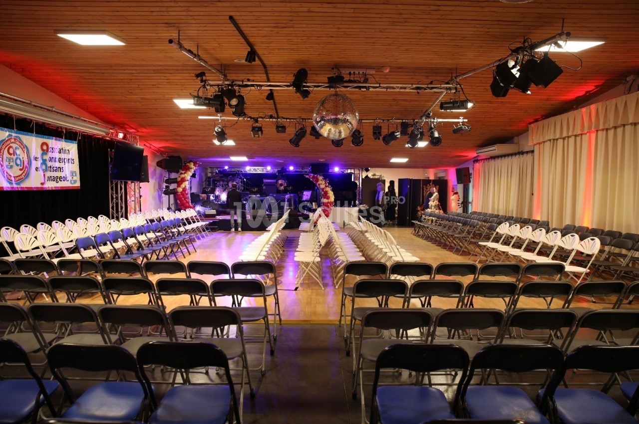 Salle aménagée pour un événement avec des rangées de chaises, une scène éclairée et une boule disco suspendue.