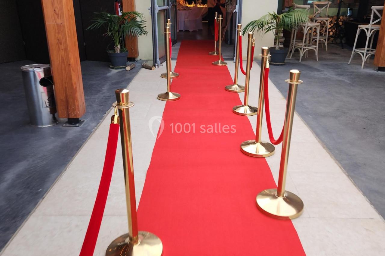 Tapis rouge bordé de poteaux dorés et cordons rouges menant à une entrée lumineuse.