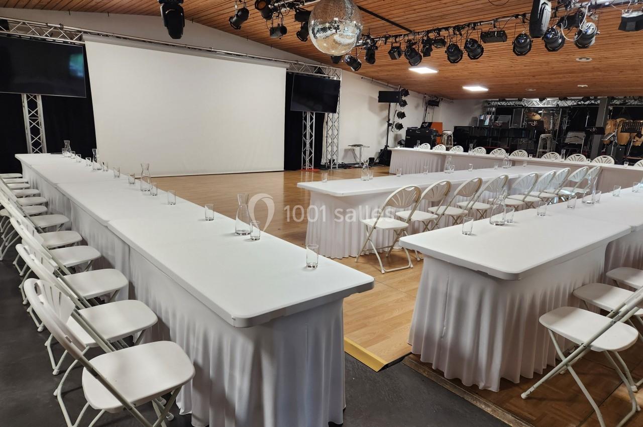 Salle de conférence aménagée avec des tables blanches en U, des chaises pliantes et un écran de projection.