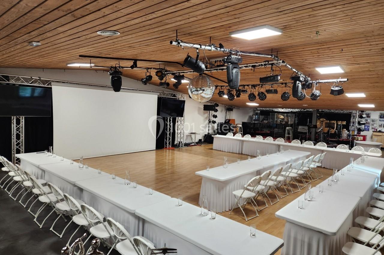 Salle de conférence avec tables en U, chaises blanches, écran de projection et éclairage suspendu.