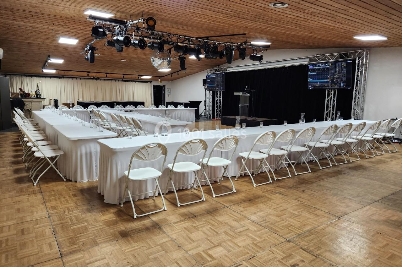 Salle de conférence avec tables disposées en U, chaises blanches pliantes et scène équipée de matériel audiovisuel.