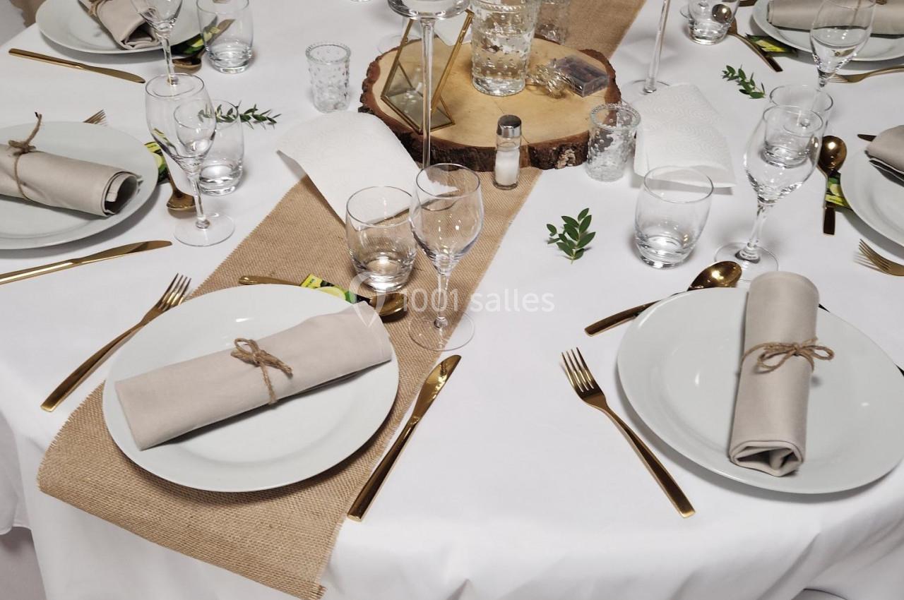 Table dressée avec nappes blanches, couverts dorés, serviettes beige nouées, chemins de table en jute et décorations…