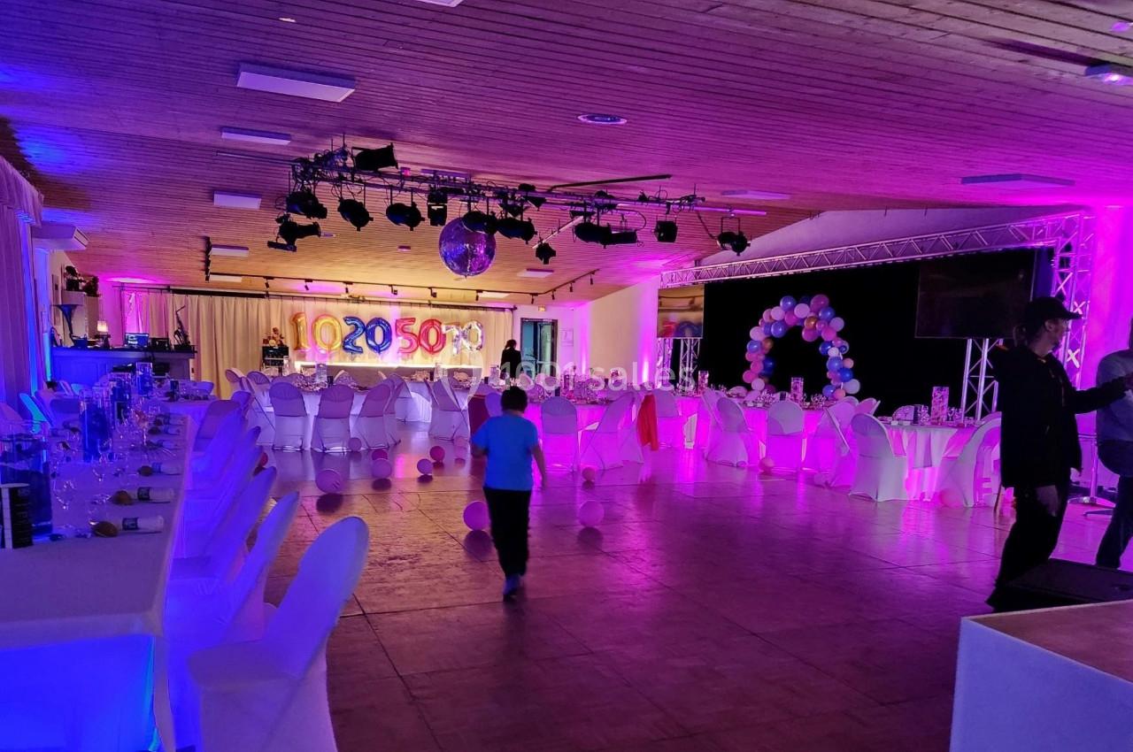 Salle de réception décorée avec des tables dressées, des ballons et des éclairages colorés, avec quelques personnes…