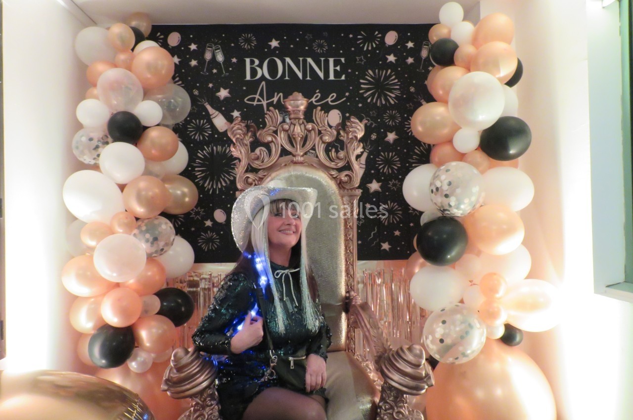 Femme souriante assise sur un trône décoré, entourée de ballons et d'une affiche ’Bonne Année’ festive en arrière-plan.