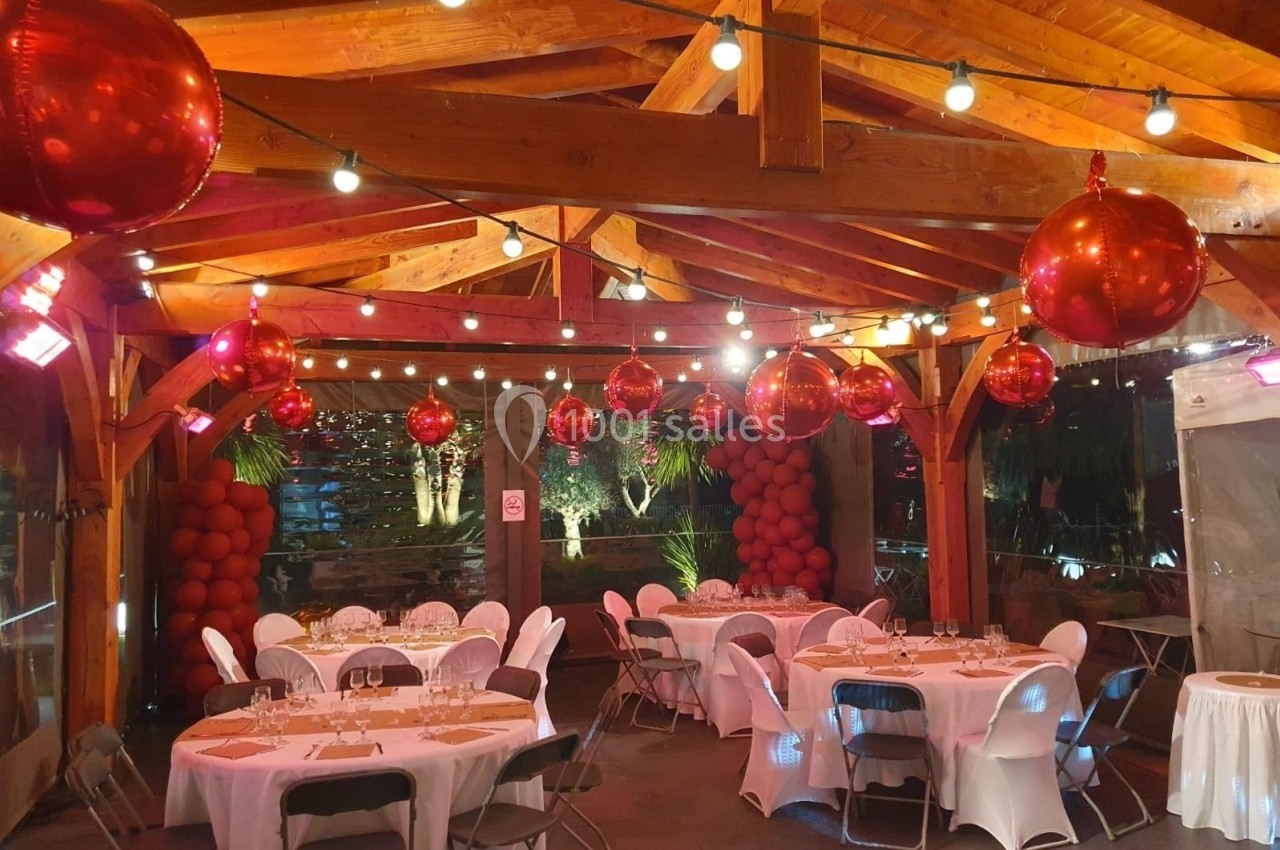 Salle décorée avec des ballons rouges et des guirlandes lumineuses, tables rondes dressées pour un événement.