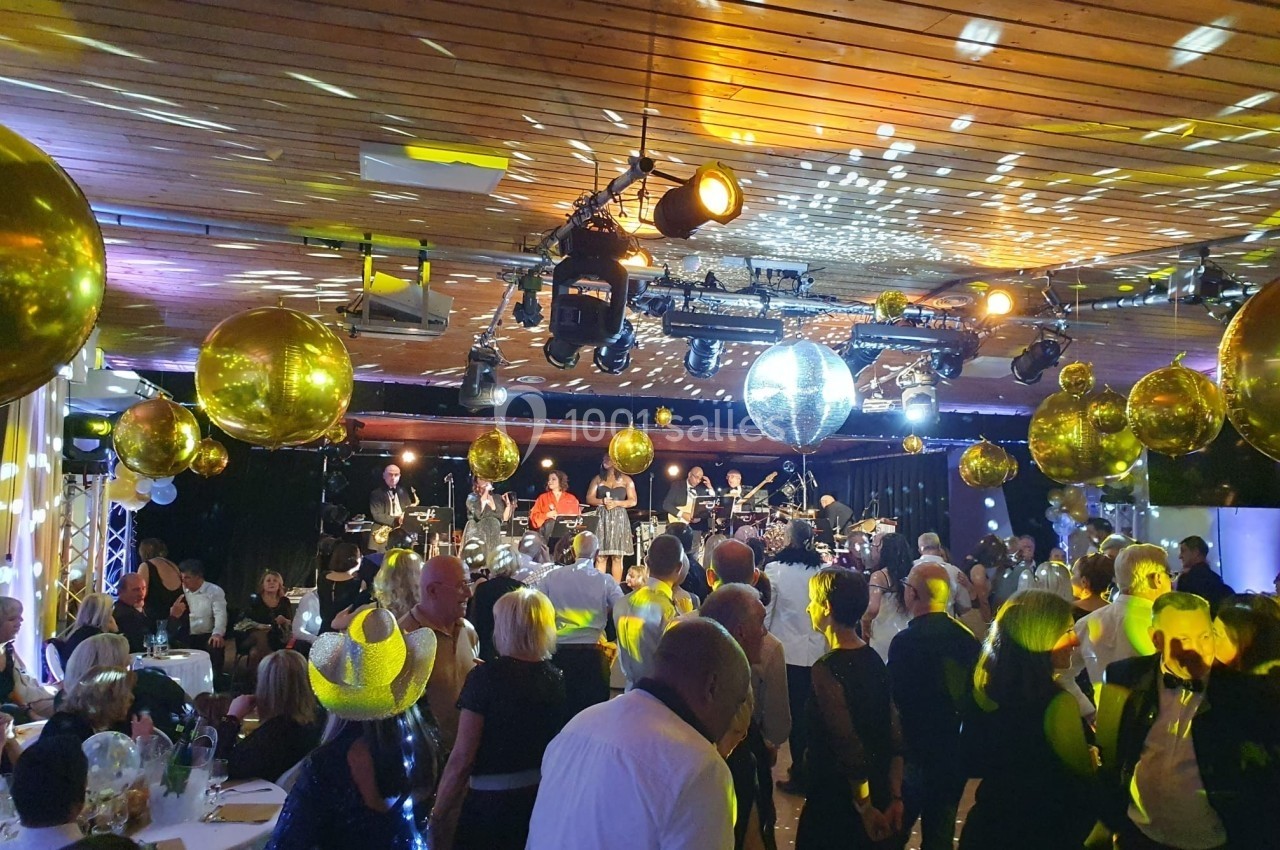 Salle festive avec des invités dansant sous des lumières colorées, des ballons dorés et une scène avec un groupe musical.