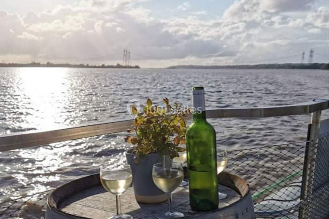 Bouteille de vin et trois verres sur un tonneau près d'une plante, avec vue sur un fleuve sous un ciel nuageux.