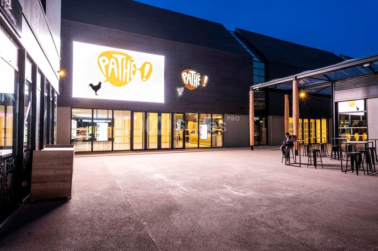 Façade d'un cinéma Pathé éclairée de nuit, avec une terrasse et quelques tables en extérieur.