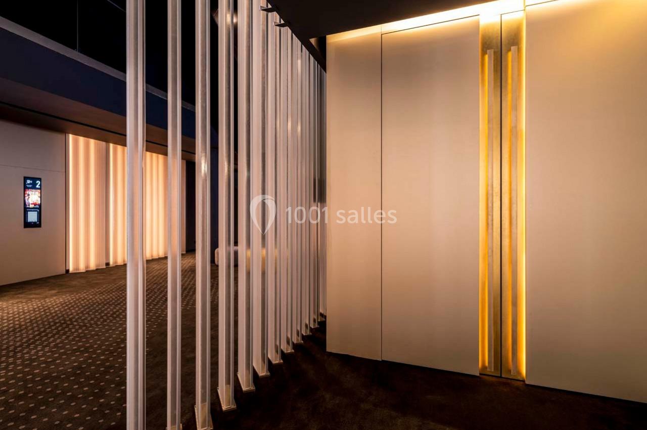 Couloir moderne avec éclairage tamisé, murs clairs et portes d'ascenseur illuminées par des bandes lumineuses verticales.