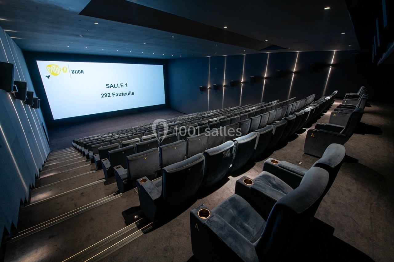 Salle de cinéma moderne avec rangées de fauteuils en velours gris et grand écran affichant ’Salle 1, 282 fauteuils’.