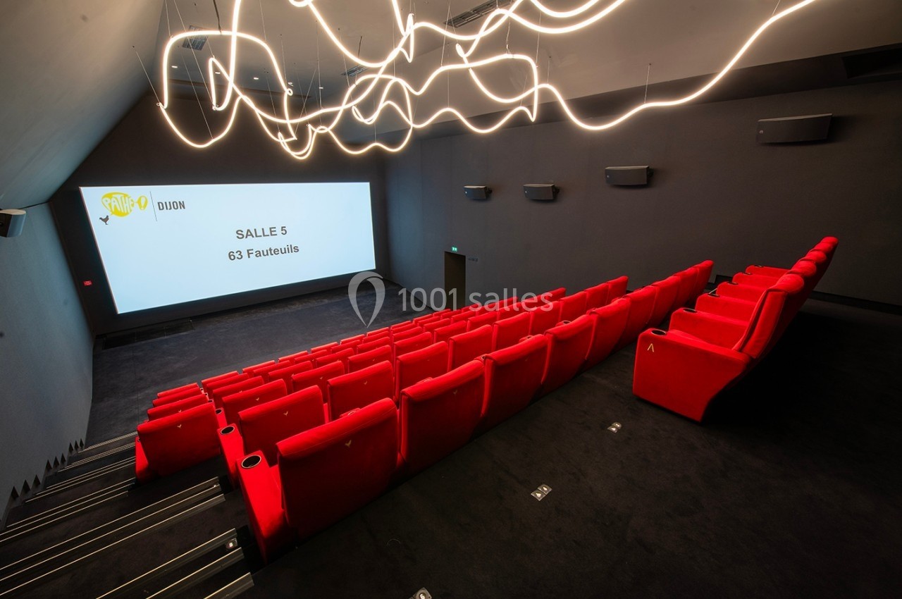 Salle de cinéma moderne avec des sièges rouges, un écran blanc affichant ’Salle 5 - 63 Fauteuils’ et un éclairage design.