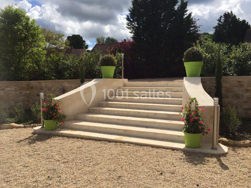 Escalier extérieur en pierre entouré de pots de fleurs vert vif, avec un jardin et un mur en pierre en arrière-plan.
