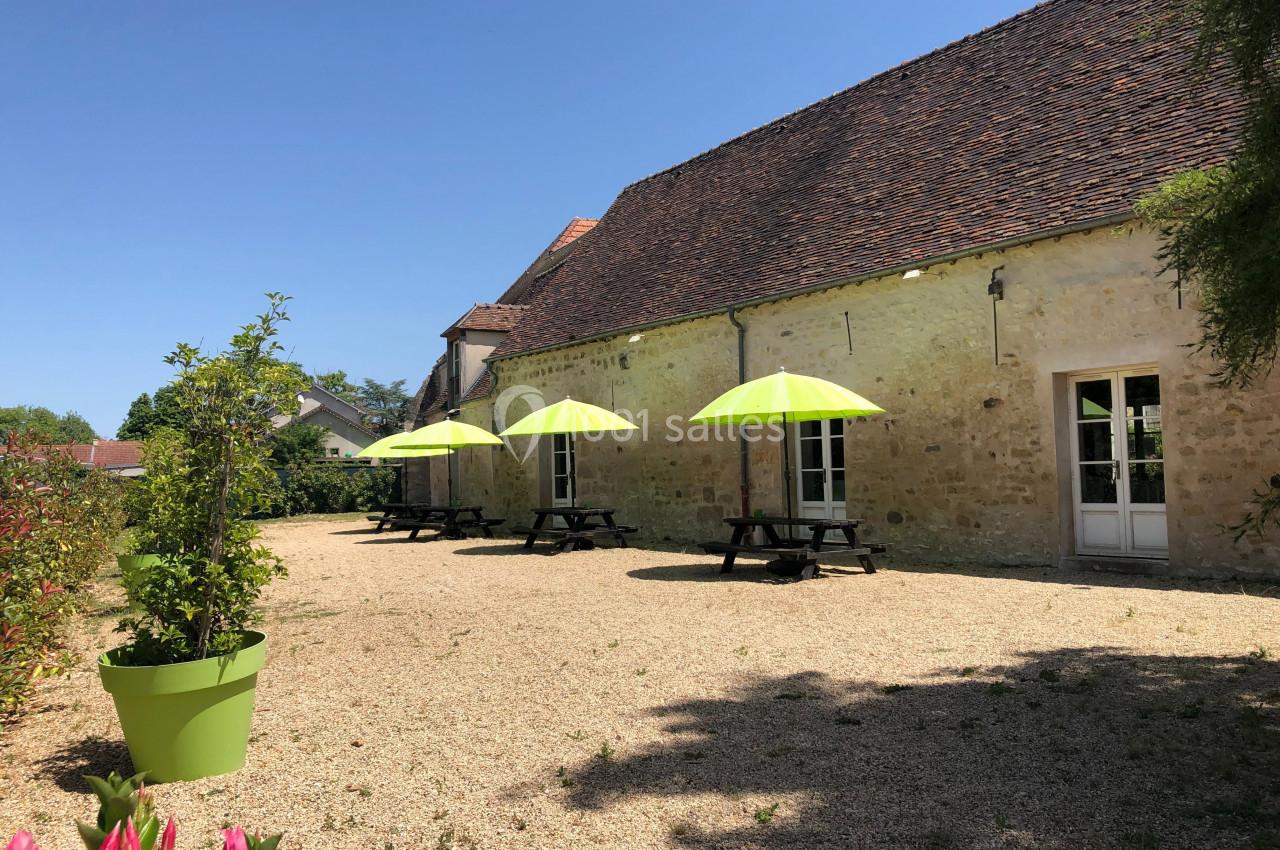 Cour extérieure avec tables en bois et parasols verts, devant un bâtiment en pierre sous un ciel dégagé.