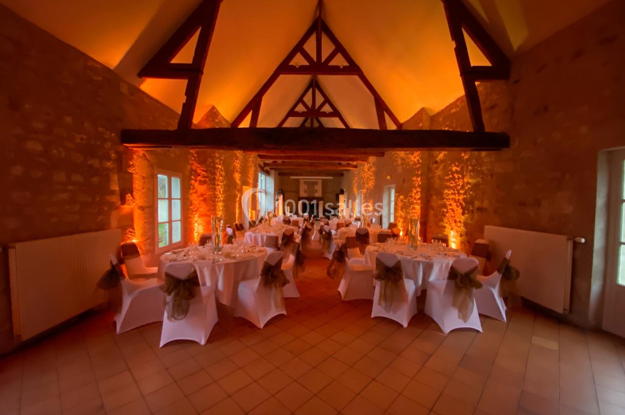 Salle de réception avec tables rondes dressées, éclairage chaleureux et poutres apparentes en bois.