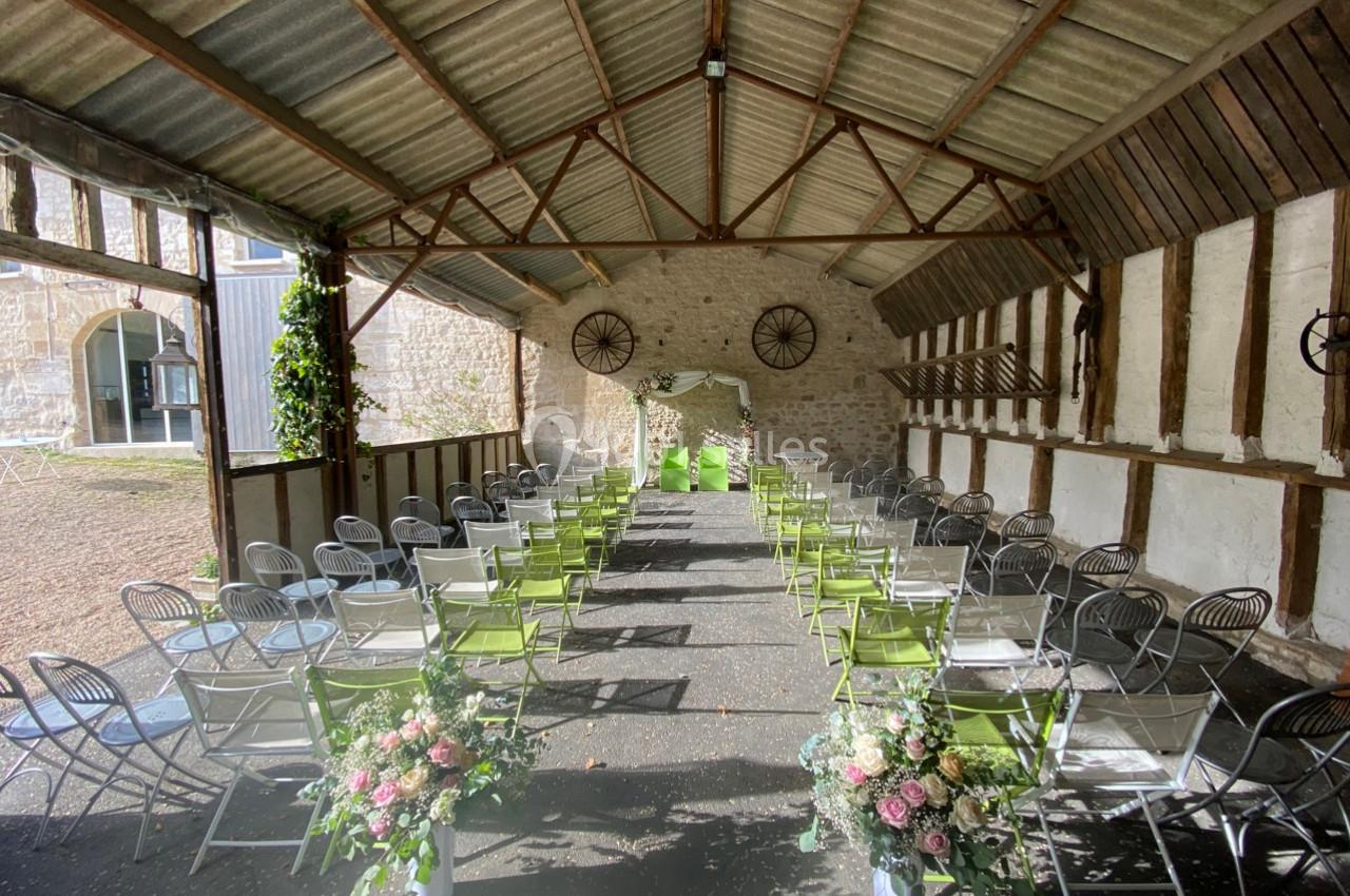 Salle de cérémonie en plein air sous un toit en tôle, avec des rangées de chaises et des décorations florales.