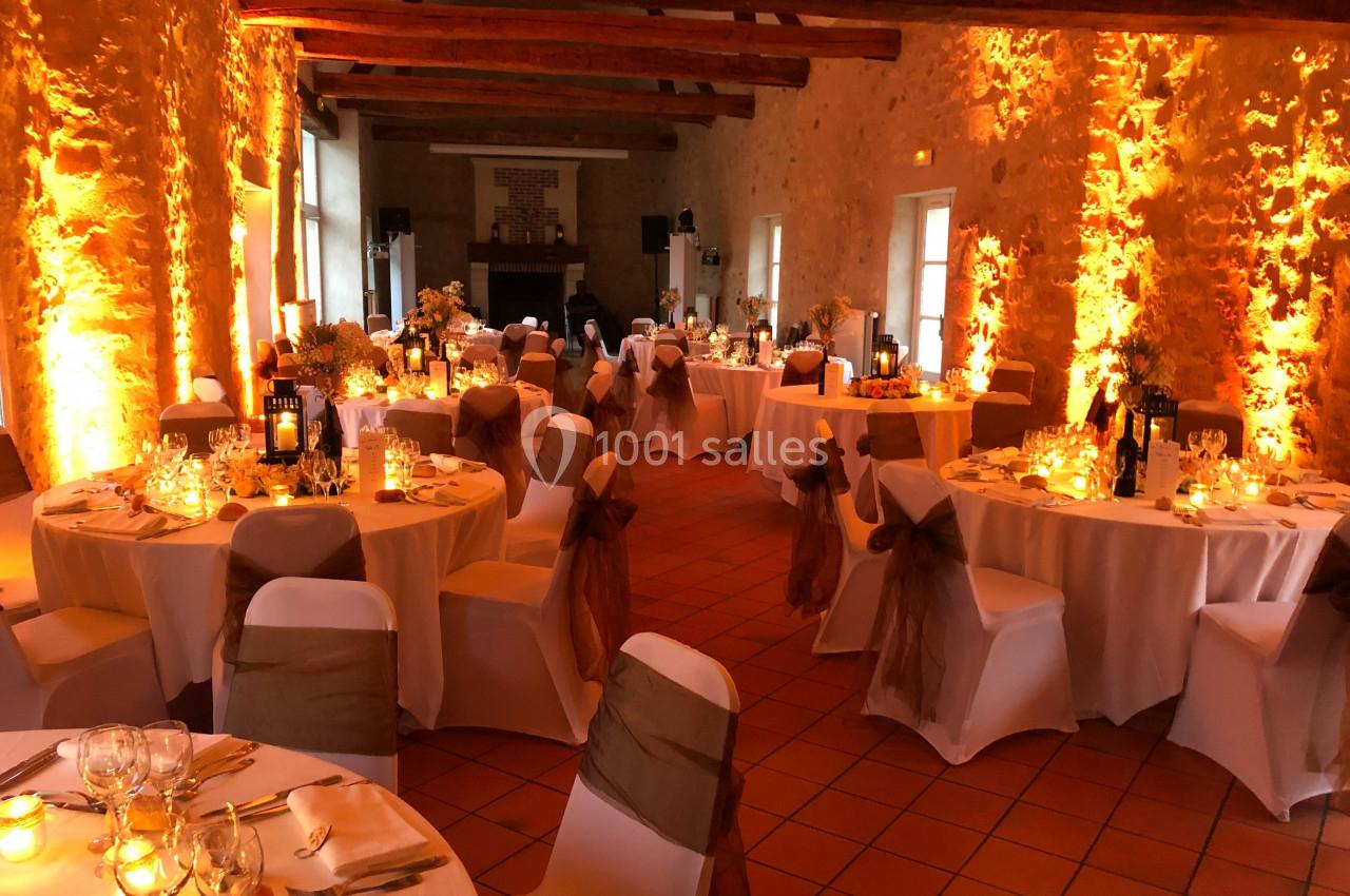 Salle de réception décorée avec des tables rondes, nappes blanches, chaises ornées de rubans et éclairage chaleureux.