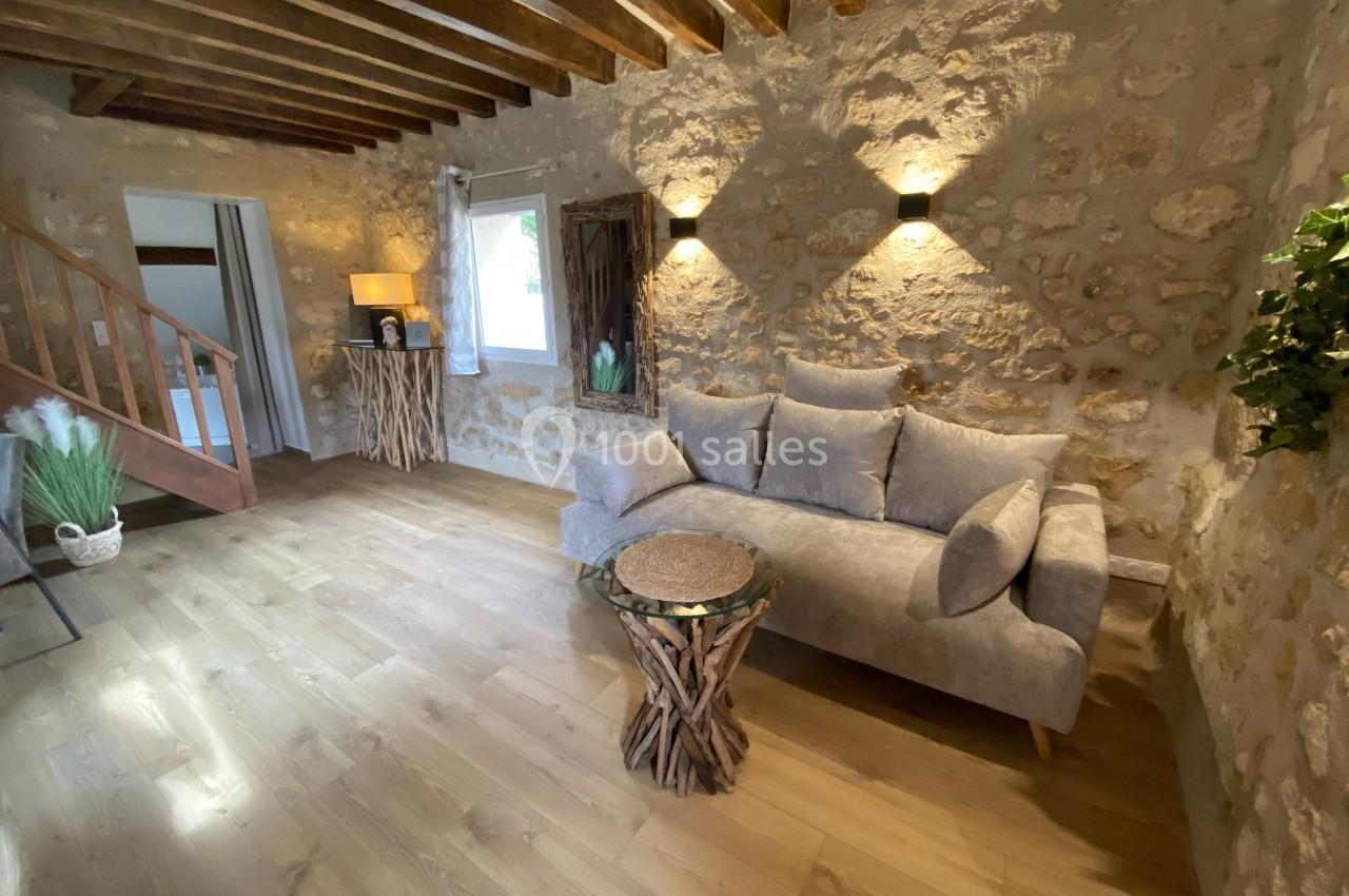 Salon chaleureux avec murs en pierre, canapé gris, table en bois naturel et escalier en bois visible à gauche.