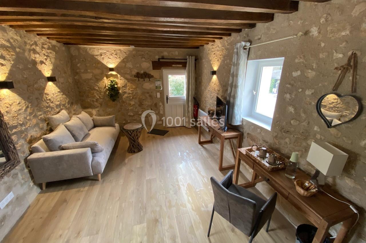 Salon chaleureux avec murs en pierre, poutres apparentes, canapé, bureau en bois et lumière naturelle.