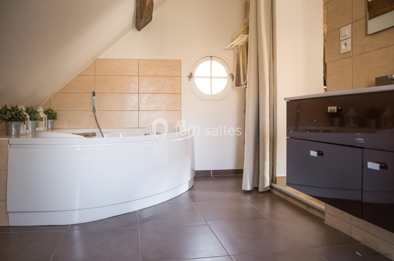 Salle de bain mansardée avec baignoire d'angle, meuble vasque brun et fenêtre ronde éclairant la pièce.