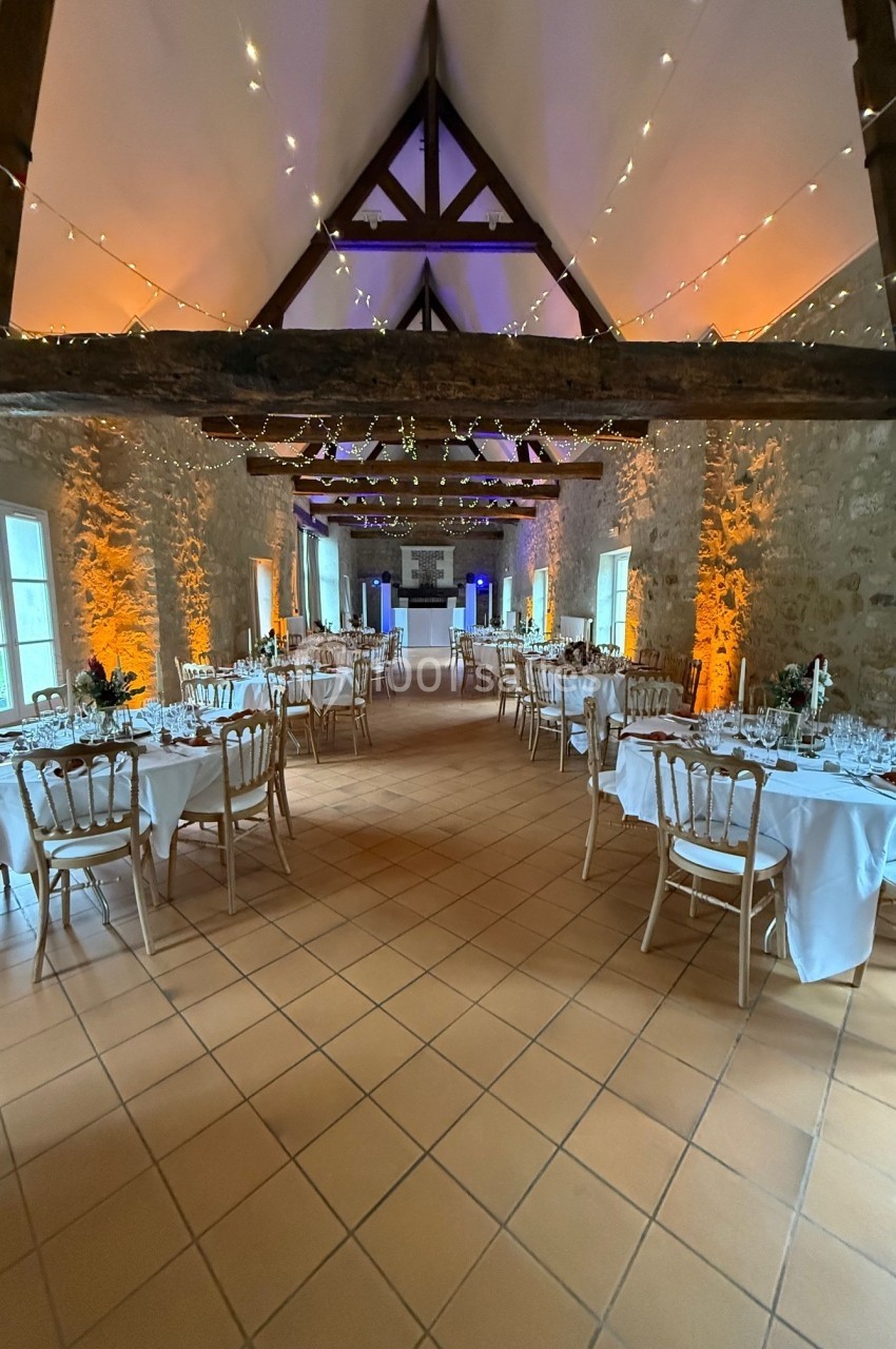 Salle de réception avec tables dressées, chaises en bois clair, murs en pierre et guirlandes lumineuses suspendues.