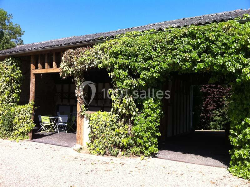 Grange en bois partiellement couverte de végétation, avec une ouverture et un espace aménagé avec des chaises.