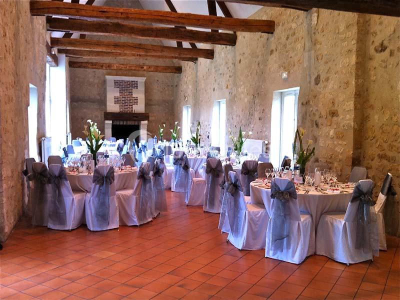 Salle de réception avec tables rondes décorées de nappes blanches, chaises ornées de nœuds gris et arrangements floraux.