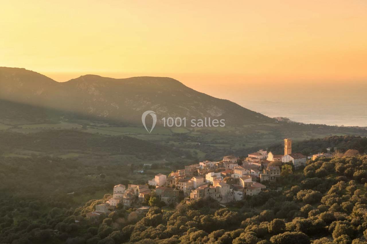 Village perché entouré de végétation, baigné par une lumière dorée au coucher du soleil, avec des collines en arrière-plan.
