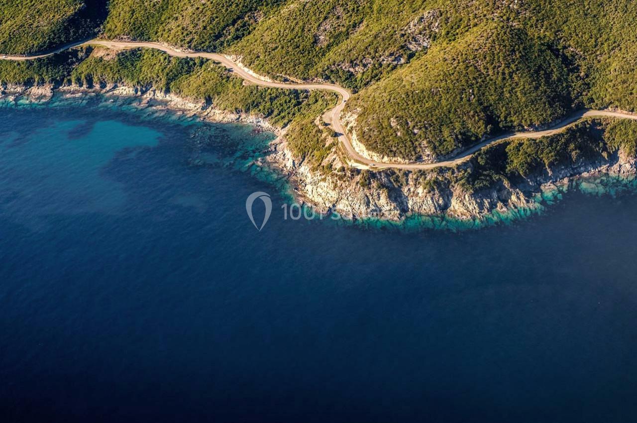 Vue aérienne d'une route sinueuse longeant une côte rocheuse bordée de végétation et d'une mer bleue.