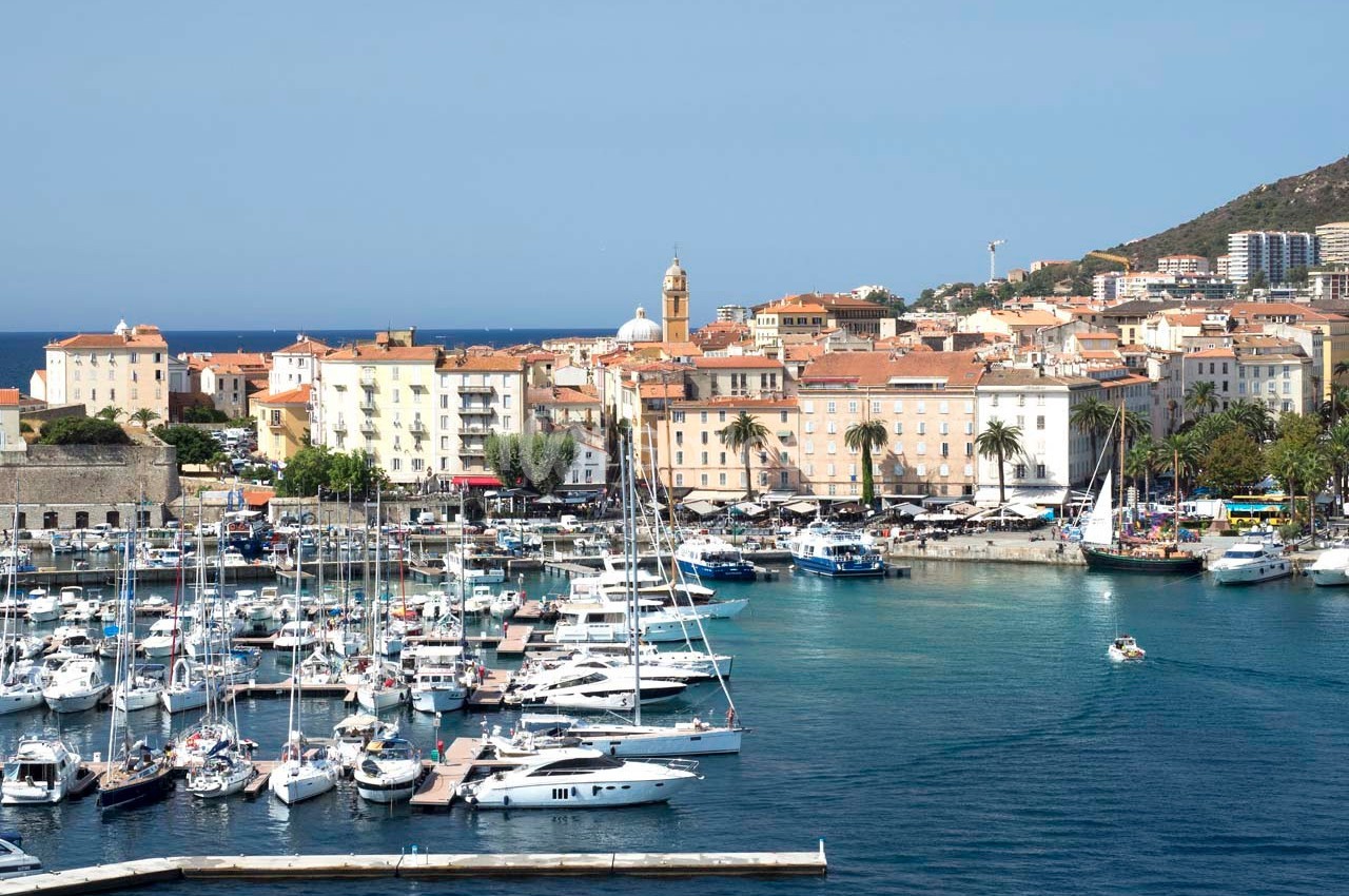 Port avec des voiliers et yachts, bordé par une ville côtière aux bâtiments clairs et collines en arrière-plan.
