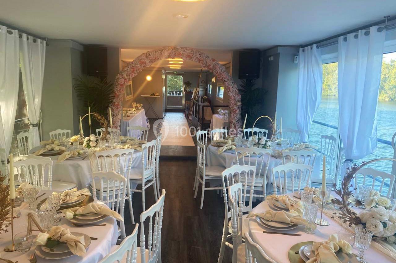 Salle de réception élégamment décorée avec des tables dressées, des chaises blanches et une arche florale, vue sur l'eau.