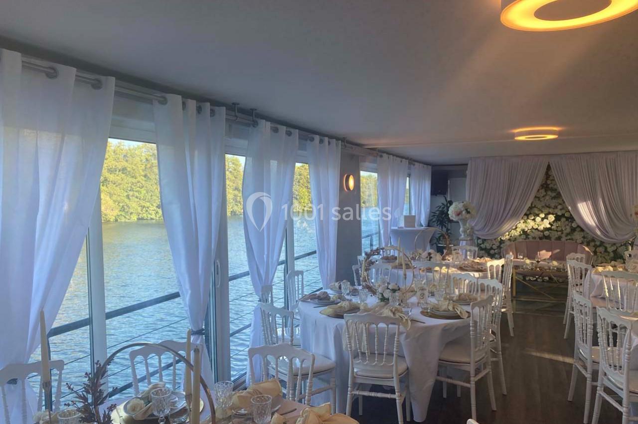 Salle de réception élégante avec vue sur l'eau, décorée de tables dressées et de fleurs blanches.