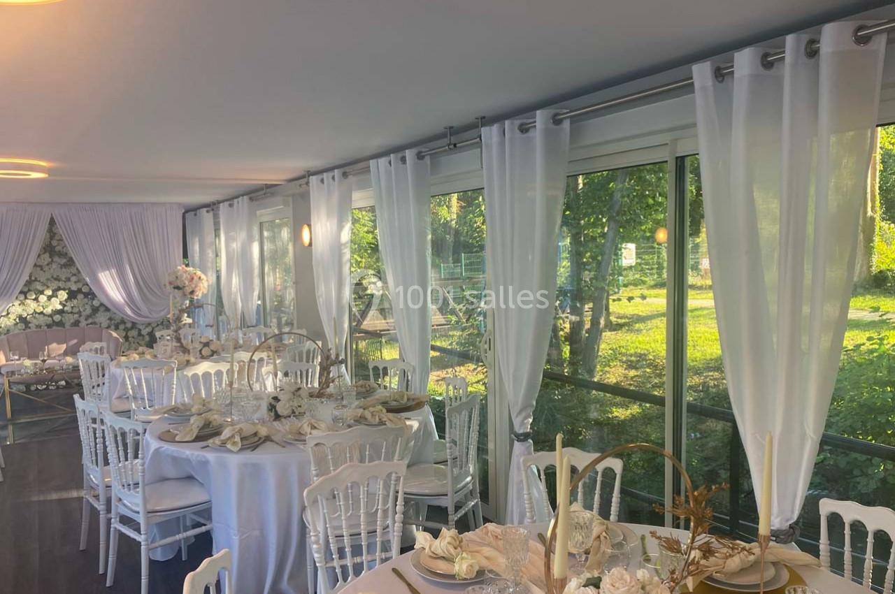 Salle de réception lumineuse avec tables décorées de nappes blanches et vue sur un jardin à travers de grandes baies vitrées.