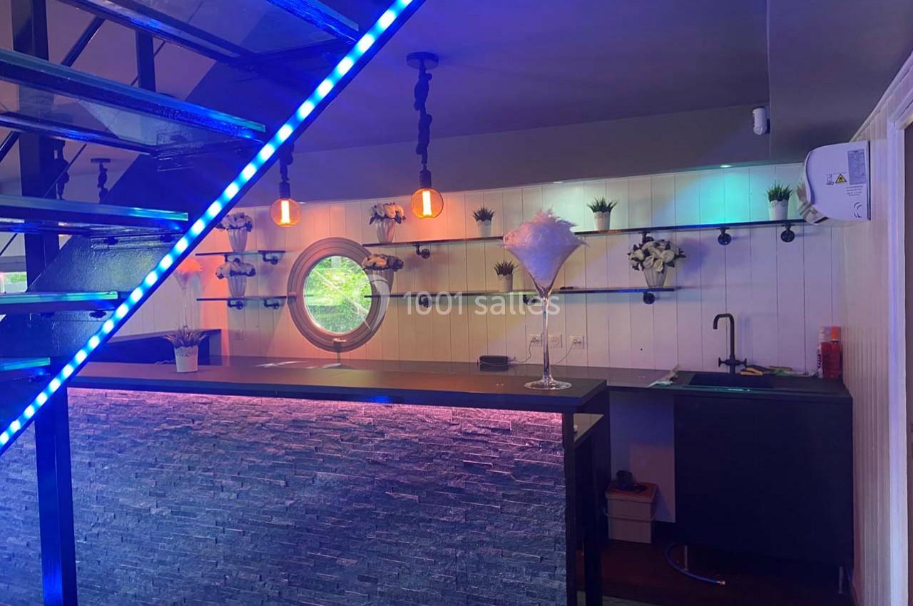 Bar moderne avec comptoir en pierre, éclairage coloré, étagères décorées et un escalier métallique en premier plan.