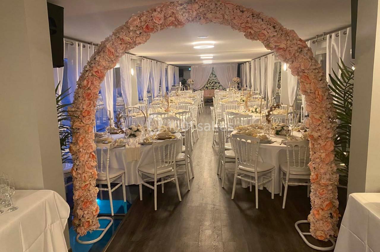 Salle de réception décorée avec des tables dressées et un arc floral rose à l'entrée.